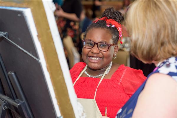 A student wearing glasses, pearl earrings and a pearl necklace smiles wide at the camera. She is standing in front of an easel. 